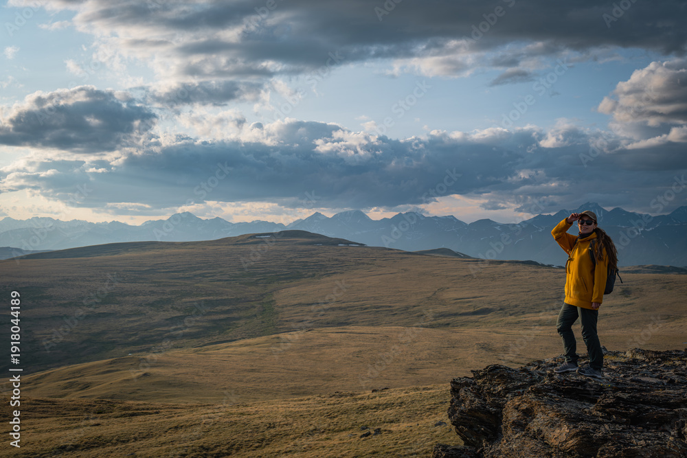 Fototapeta premium Female hiker in yellow jacket standing on edge of rocky cliff looking at vast mountain plateau under cloudy sky during sunset travel adventure. 