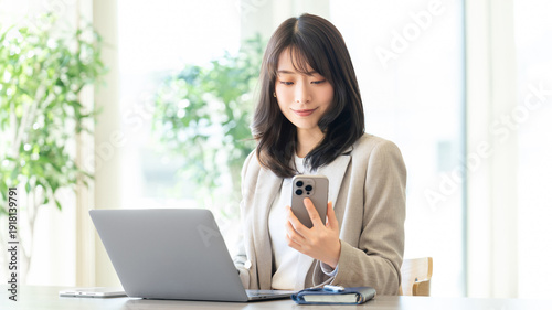 Foto ノートパソコンを前にスマートフォンを確認する女性ビジネスパーソン, Businesswoman checking smartphone in front of