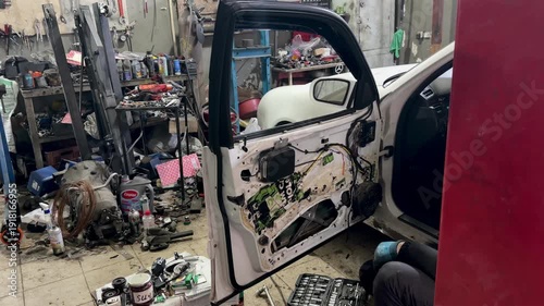 Auto mechanic working on a car door repair in a professional garage. Close-up of disassembled white car door with internal mechanisms, wiring, and tools visible in a workshop background.