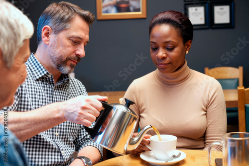 Adult man pouring tea into cup for adult Black woman sitting at table with senior woman, group engaging in social interaction in casual setting
