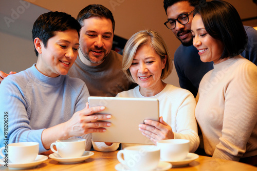 Group of adult woman, adult man, young Black man, young woman and young man gathering around tablet, smiling and looking at screen