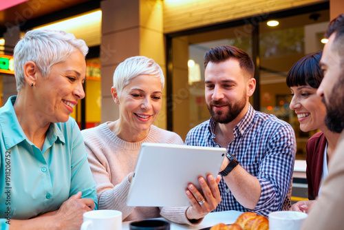 Group of adult people sitting together smiling and looking at tablet screen while having coffee and pastries at outdoor cafe table