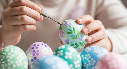 Child's hands delicately paint intricate floral patterns onto a pale green Easter egg using a fine-tipped brush above a collection of decorated eggs.