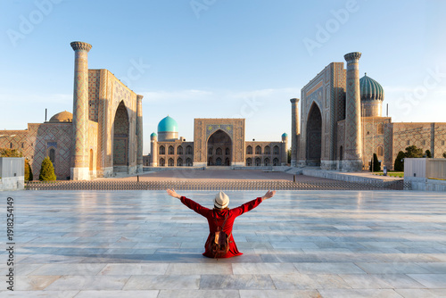 Uzbekistan, Samarcanda - young girl sitting with open arms looking Registan square. Translation: In the name of Allah Almighty who creates
