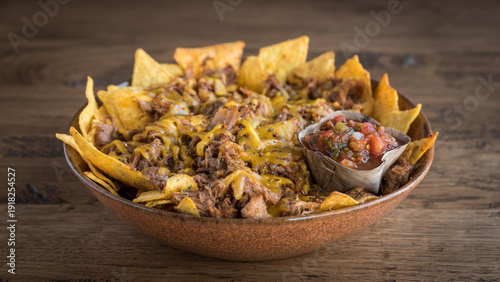 Close up of nachos loaded with pulled pork and cheese with a salsa dip in a bowl on a wooden table