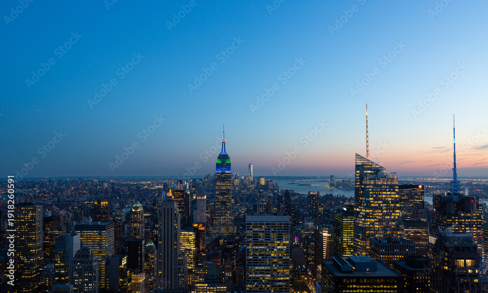 Obraz premium Panoramic view of New York City skyline at dusk featuring illuminated skyscrapers and iconic architecture under a colorful sunset sky. Urban cityscape, travel and business concept.