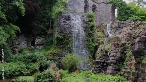 Waterfall Closeup Over Mossy Stone Arch With Vibrant Yellow Flowers And Lush Greenery Powerful Spray Drifts Across Camera, Textured Stone And Fern Fronds Frame Foreground. Kassel, Germany.