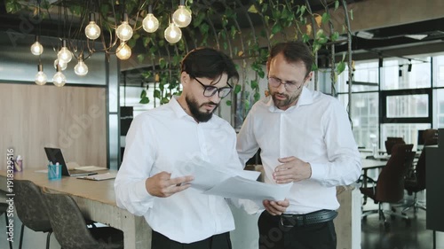 Two men discuss papers in a modern office space with hanging lights and a plant wall during daylight hours