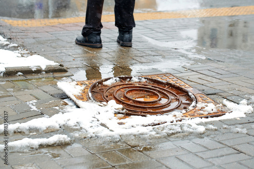 Dangerous rusted manhole cover protruding from grey brick sidewalk in the city during winter snow removal work, creating serious tripping hazard for pedestrians near slushy snow and deep puddles
