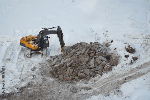 Canvas Print A yellow excavator operates amidst snow and a large pile of concrete rubble at a winter construction site