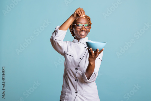 African american professional cook happily sprinkling pinch of salt into bowl to enhance flavor. Happy female chef adds final touches to freshly prepared meal, showing joy and passion for cooking.