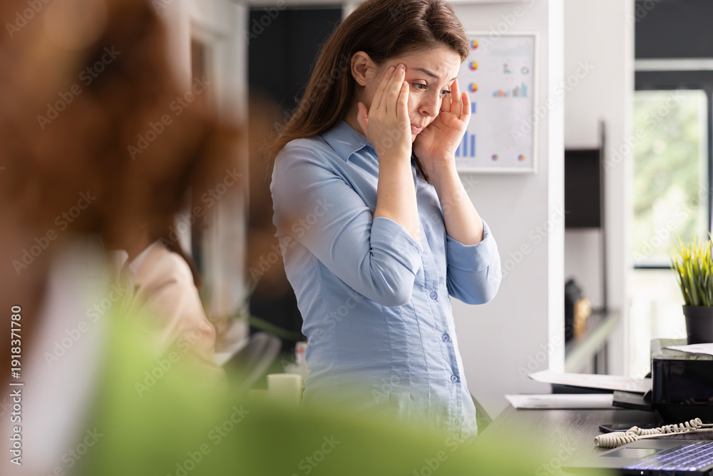 Fototapeta premium Stressed professional woman touches head in frustration and analyzes marketing strategy files on digital device. Tired employee experiences pressure and anxiety in busy workplace.