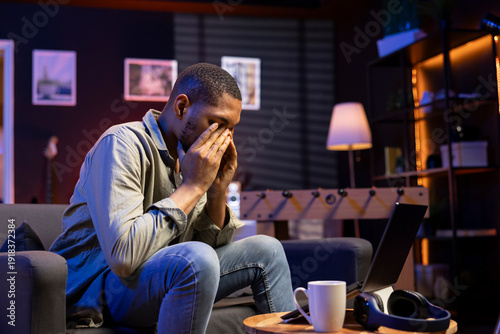 Young black person overworked with a headache in living room, working remotely under stress and pressure from his boss. Young adult frustrated and worried, struggling alone with tasks.