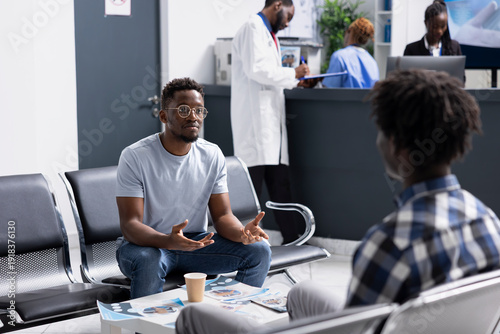 Φωτογραφία Two male clients sit together in hospital waiting room, speaking before scheduled medical checkups
