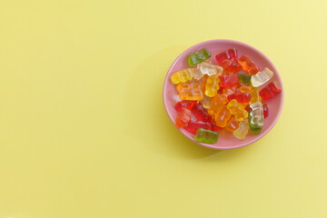 A pink ceramic bowl filled with a generous assortment of colorful gummy bear candies in red, orange, yellow, green, and white is photographed from above against a bright yellow-green background.