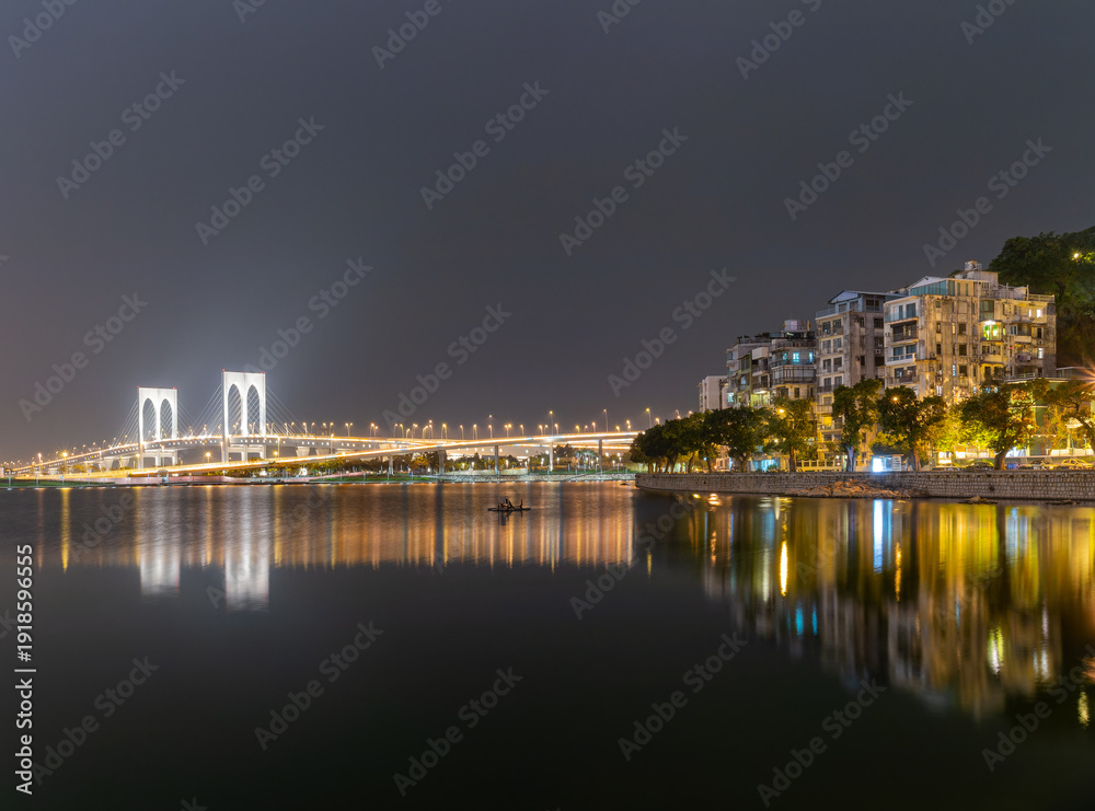 Fototapeta premium Illuminated bridge and cityscape reflected in calm water at night.