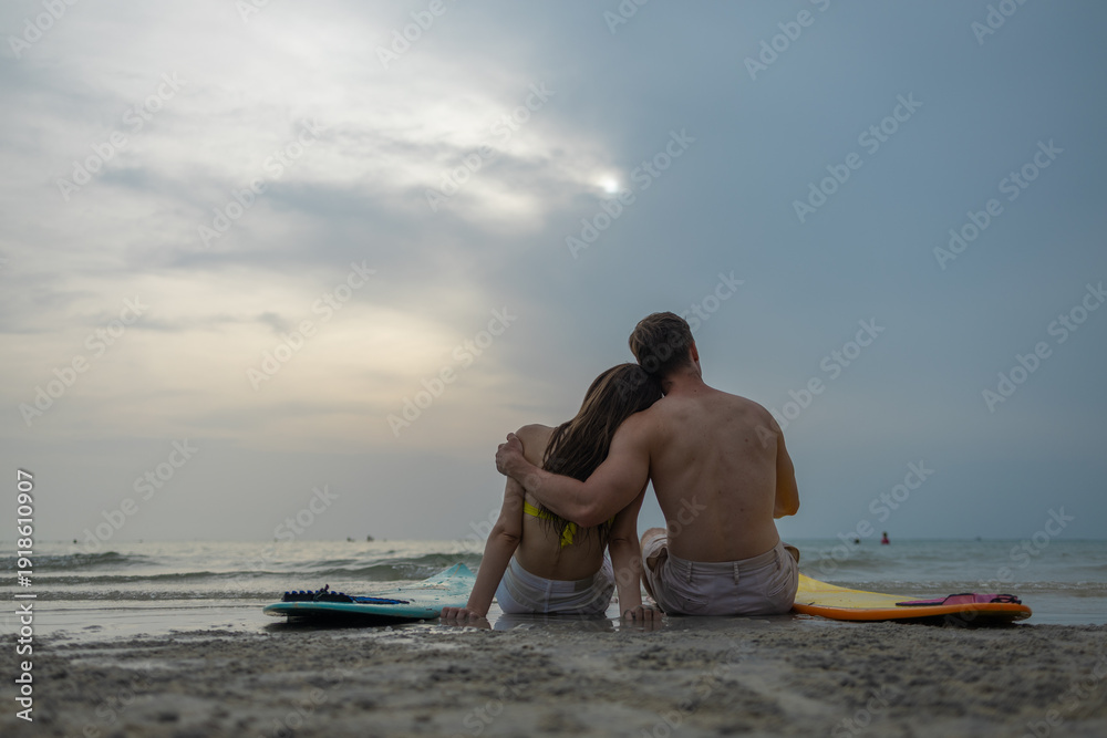 Obraz premium Romantic couple sitting on beach with surfboards at sunset, embracing by the ocean. Summer vacation, love relationship, tropical travel lifestyle and peaceful seaside moment concept.