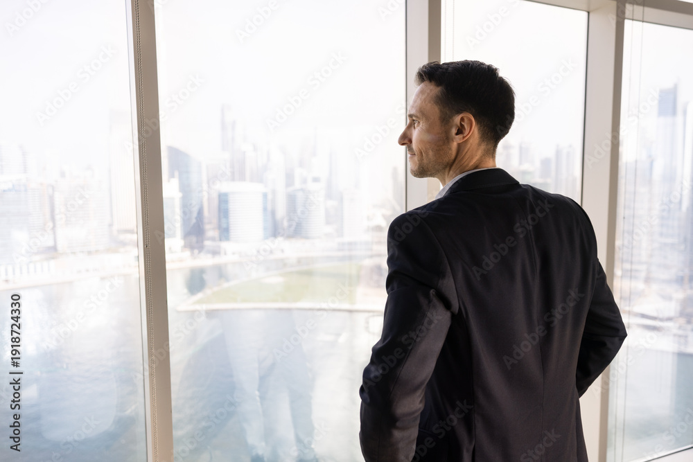 Fototapeta premium Positive thoughtful young property investor man looking at city view out of office window, thinking on real estate project, planning investment, enjoying work break, leisure, daydreaming