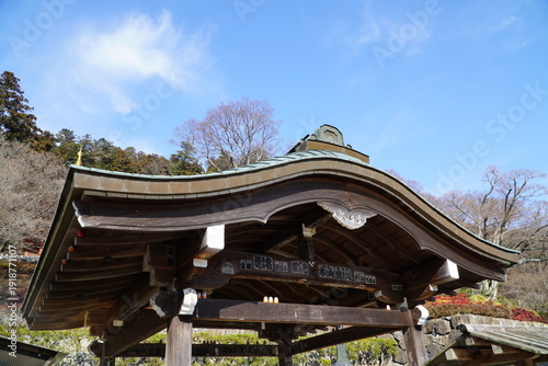 Traditional Japanese wooden pavilion roof with elegant curved architecture and intricate carvings, set against a clear blue sky and lush mountain forest. This serene structure features historical craf