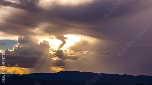 Dramatic sunset with brilliant golden sunlight piercing through the layered dark storm clouds casting light rays over the mountain range below