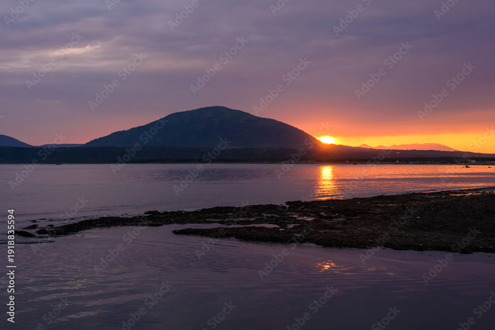 Obraz premium Golden sunlight shines across calm waters as it sets behind majestic mountains in Russia. The scene captures the beauty of nature at dusk, reflecting peace and tranquility.