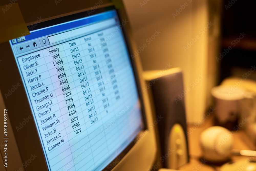 Obraz premium Close up showing computer monitor displaying employee salary spreadsheet with names, salaries, dates and bonus columns, office desk objects including baseball and speaker visible in background