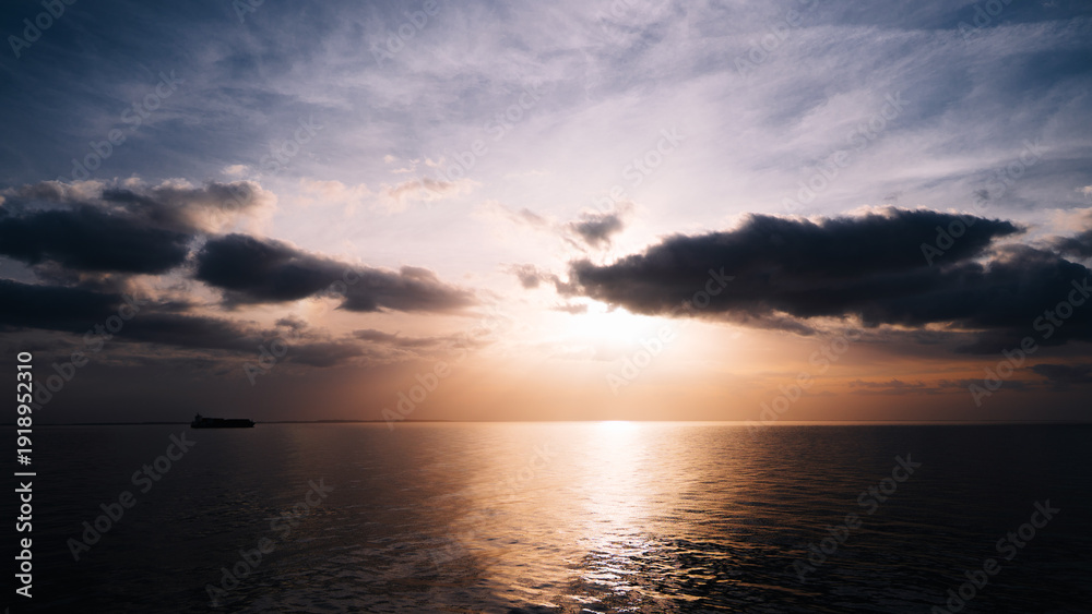 Fototapeta premium Silhouette of a cargo vessel on the horizon during sunset over calm sea. Dramatic clouds illuminated by warm evening light with golden reflection on the water surface. Peaceful maritime landscape
