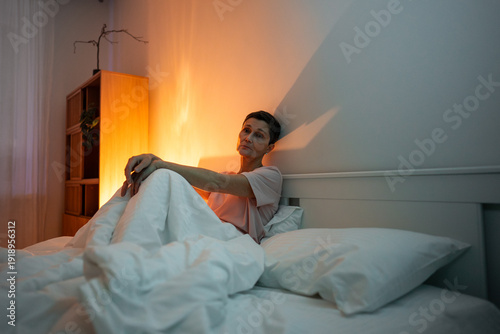Fotografia Exhausted pondering senior woman with insomnia sitting in bed in bedroom with apathy and tired expression, thinking and staring at emptiness