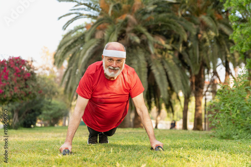 Cuadro en lienzo Sportive senior man in red shirt and headband doing push-ups on handles in outdo