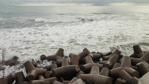 Concrete tetrapods forming a breakwater along a stormy ocean coastline with crashing waves under an overcast sky.