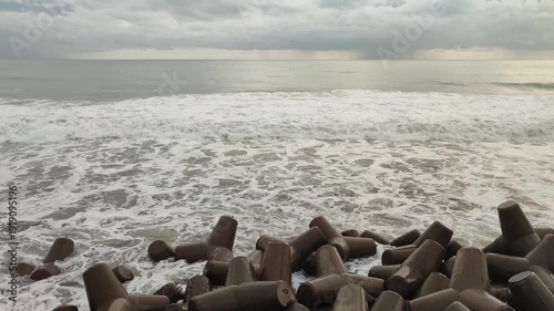 Rough sea with waves crashing against concrete tetrapods under a cloudy sky, protecting the coastline.