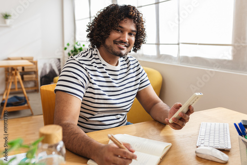 Obraz na plátne Portrait of young adult happy Indian student man smiling at camera while using smartphone at home desk