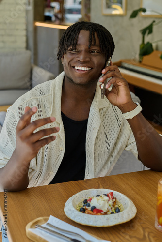 Tablou pe pânză A joyful young man smiles and chats on the phone, savoring a delicious dish in a charming cafe