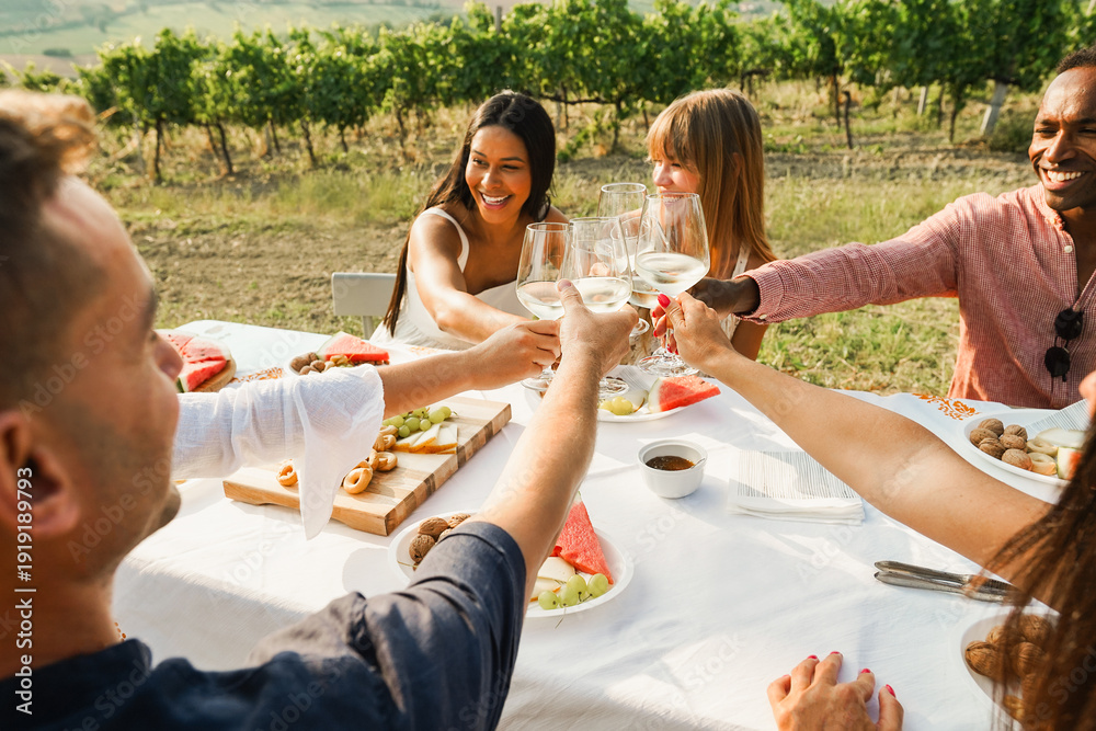 Fototapeta premium Happy adult friends having fun cheering with white wine together - Multiracial people doing party at summer time in countryside resort with vineyard in background - Main focus on hands
