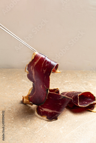 A slice of Spanish cured beef cecina delicately held with metal tweezers, lifted to the light to reveal its translucent texture and marbling. Premium artisanal charcuterie presented on a natural limes