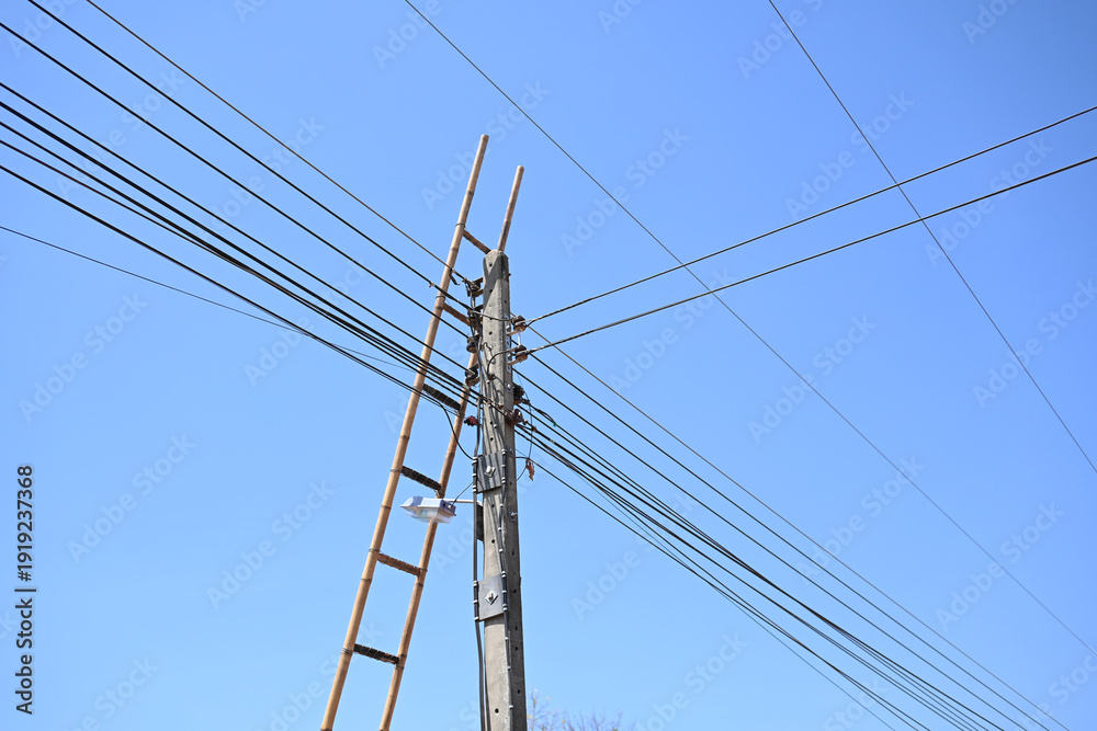 Fototapeta premium Utility pole electric wire wooden ladder blue sky power line cable network construction tool outdoor structure maintenance work minimal scene utility pole and electric wire with wooden ladder