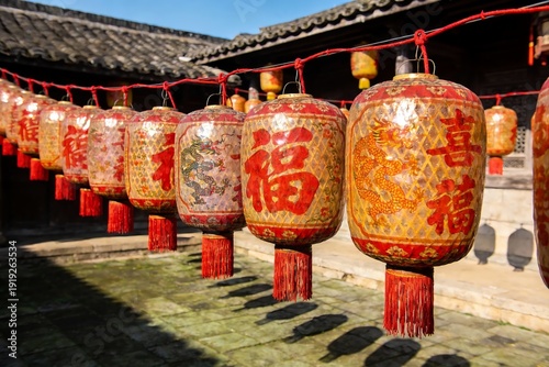 Traditional Chinese lanterns with red tassels and golden patterns hang in a row outside a historic building on a sunny day.