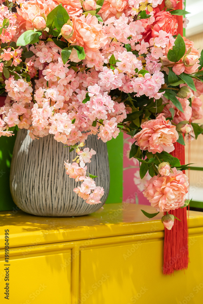 Φωτογραφία A lush pink flower bouquet rests in a striped, textured vase atop a glossy yellow cabinet, creating a vibrant centerpiece perfect for cheerful home decor, events, or festive occasions