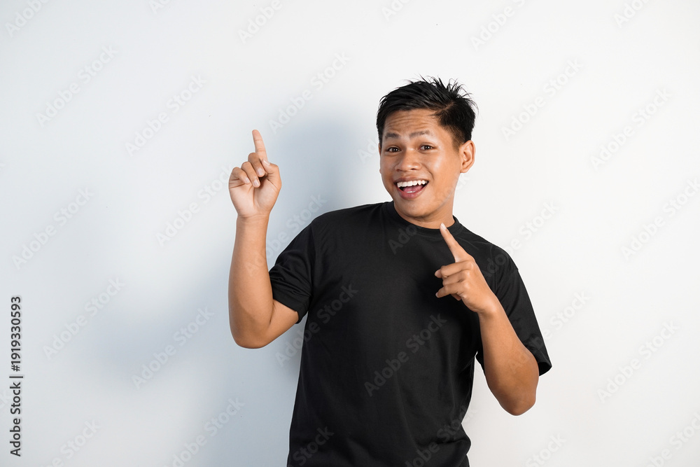 Εκτύπωση καμβά Smiling young Asian man in a black shirt points upward with both hands, conveying excitement, enthusiasm and friendly energy against a clean white background