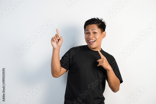 Bilde på lerret Smiling young Asian man in a black shirt points upward with both hands, conveying excitement, enthusiasm and friendly energy against a clean white background