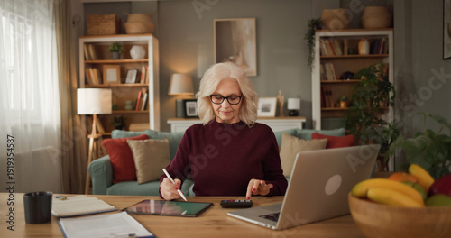 Photos In a cozy living room, a senior woman sits at a table, using a tablet for financial planning while a laptop and notebook are nearby