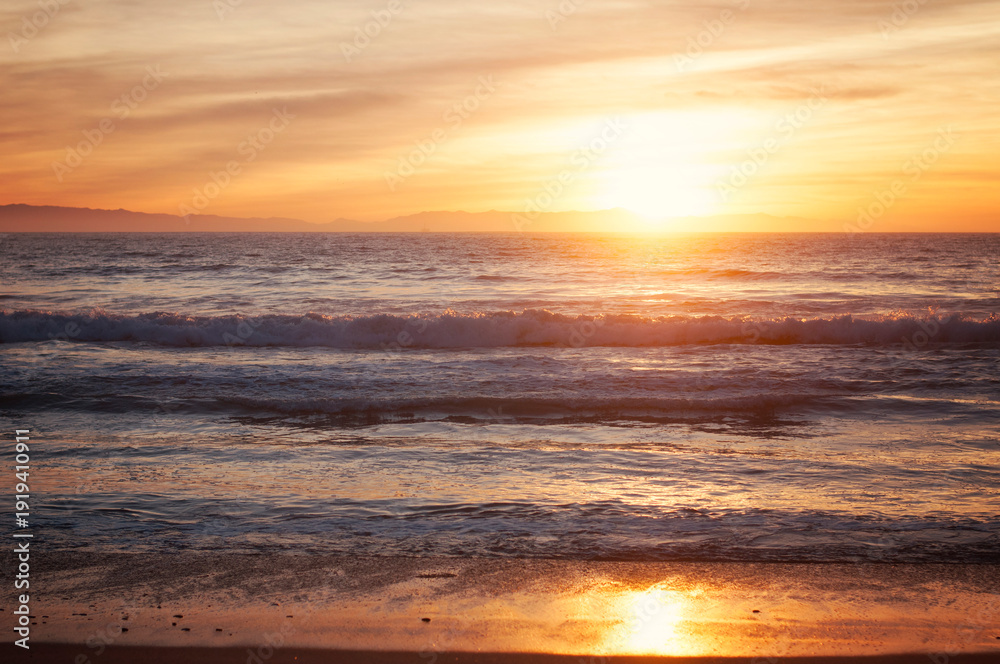 Fototapeta premium Golden sunset over ocean waves with warm orange sky reflecting on wet sand, creating a peaceful coastal landscape scene with soft light and tranquil atmosphere.