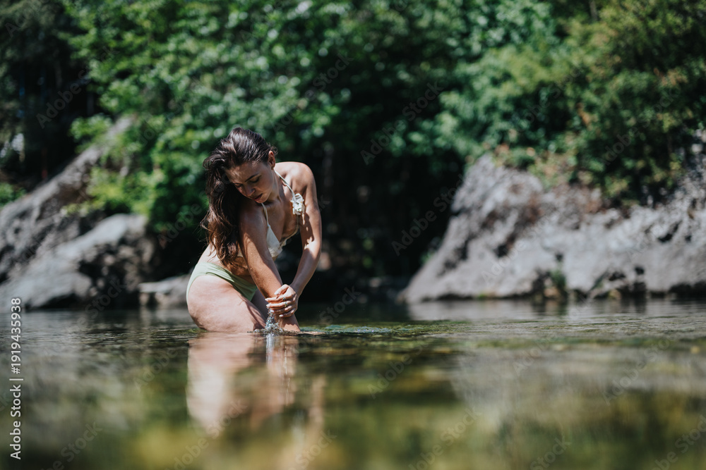 Fototapeta premium A woman in a bikini wades in a shallow river, her hands touching the water. The scene shows a peaceful summer moment by rocks and lush green trees.