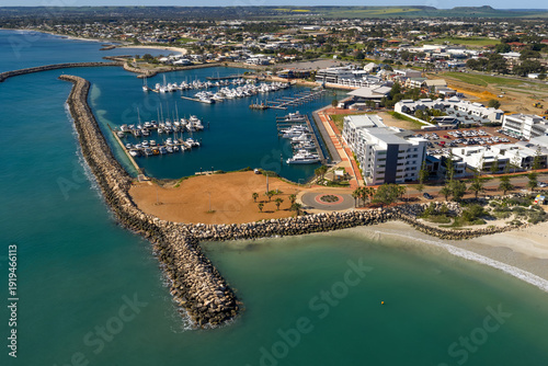 An elevated aerial view of Geraldton marina shows boats moored within a protected harbour, surrounded by coastal buildings, breakwaters, and turquoise water.