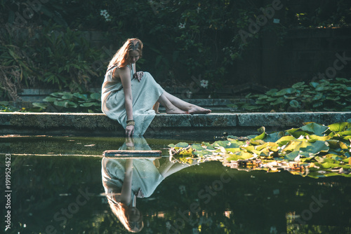 Bild auf Leinwand Thoughtful woman in a linen dress sitting on a stone edge, touching the water in a quiet garden pond at sunset