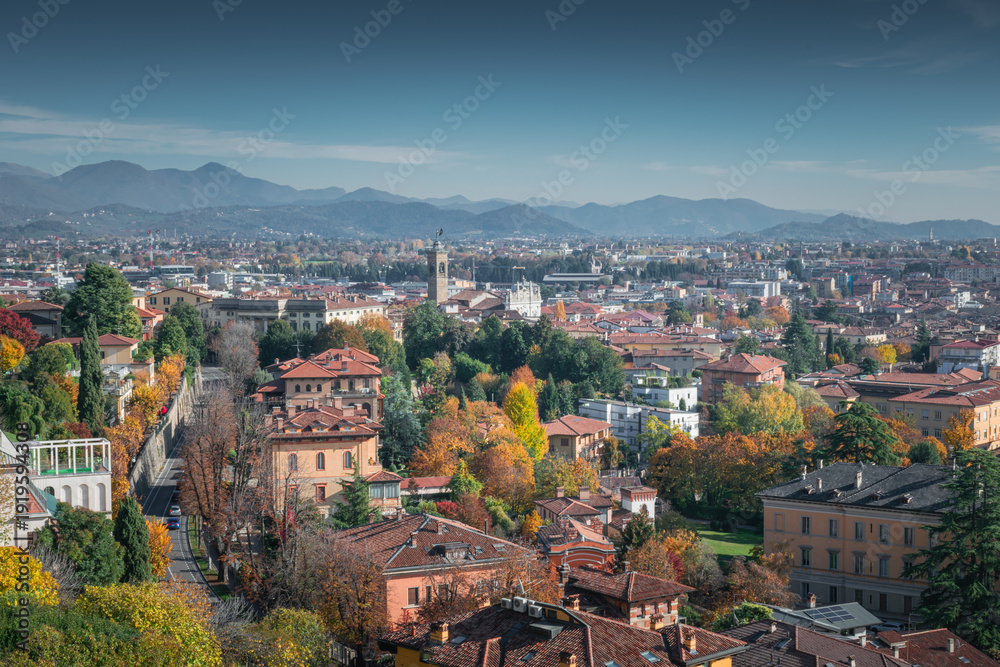 Fototapeta premium Autumn hillside city with winding road and statue‑topped clock tower facing distant mountains