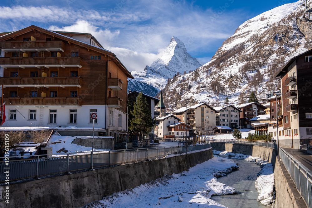Fototapeta premium Zermatt, Switzerland, shows a canal with snow banks, chalet buildings, a church steeple, and the Matterhorn. Midday light illuminates people on the quiet street.