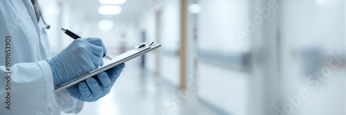 A doctor's hands hold a clipboard and write with a pen in a hospital corridor.