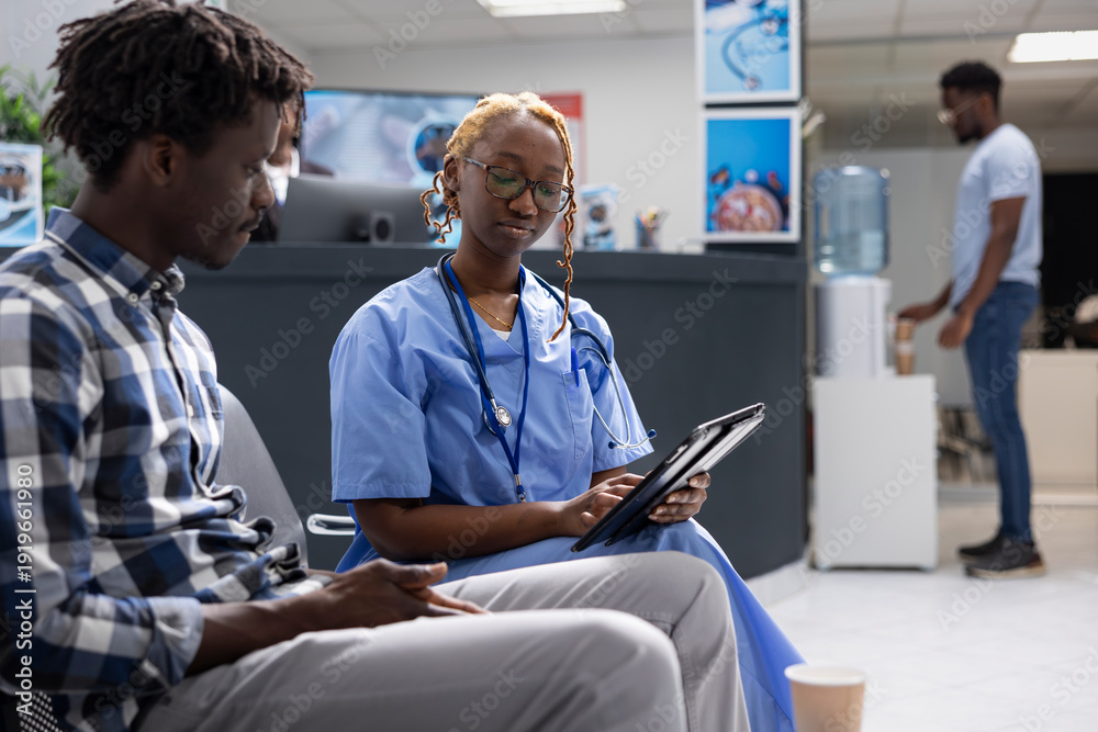 Obraz premium Nurse with glasses, uses tablet to inform young male adult about clinic procedures. Black woman in blue scrubs sitting with patient in hospital lobby, explaining diagnosis results and treatment plan.