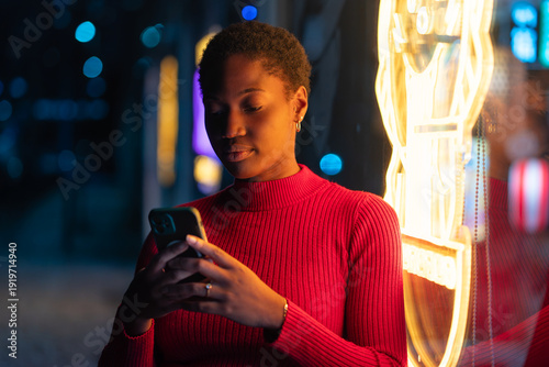 Young black woman using smartphone beside glowing neon lights on city street at night. Female in red sweater text messaging on mobile phone